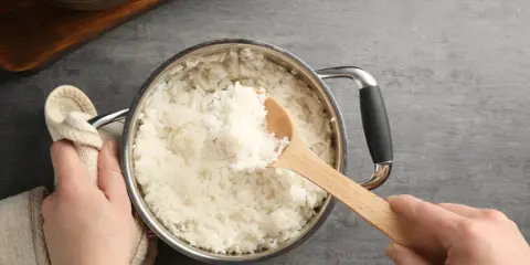 Hände halten einen Topf mit frisch gekochtem Reis. Eine Holzschaufel hebt eine Portion Reis heraus. Die Frage "wie lange kocht man reis" wird hier visuell beantwortet.