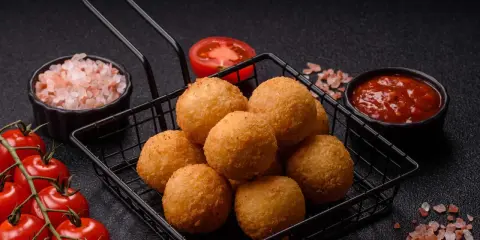 Goldbraune Arancini im Korb, bereit für ein köstliches arancini rezept. Dazu frische Tomaten, Salz und Dip.