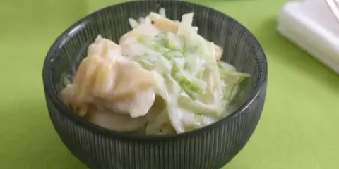 Ein cremiger Kartoffelsalat mit Joghurt und knackigem Gemüse, serviert in einer dunklen Schüssel auf einem hellgrünen Tischtuch.