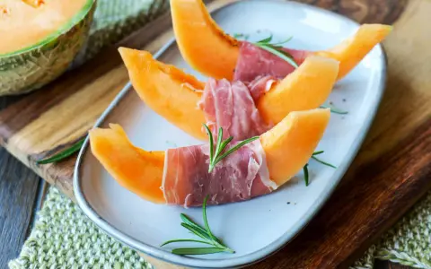 Melone mit Schinken, garniert mit Rosmarin, auf einem Teller angerichtet. Ein halber Melone liegt daneben auf einem Holzbrett.