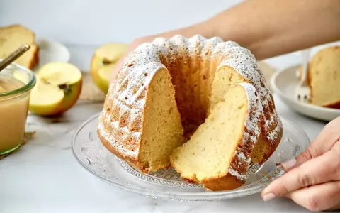 Saftiger Apfelkuchen mit Puderzucker, daneben ein Glas Apfelmus und frische Äpfel.