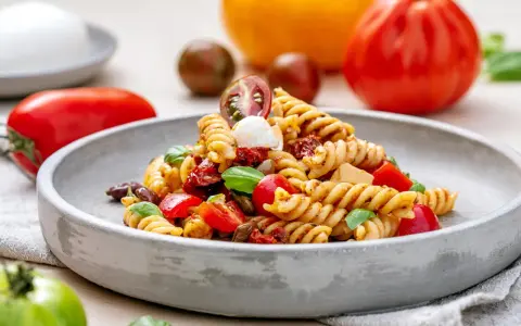 Ein köstlicher italienischer Nudelsalat mit Fusilli, Tomaten, Mozzarella und Basilikum auf einem grauen Teller, umgeben von frischen Tomaten.