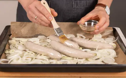 Bratwurst im Backofen: Zwiebelringe auf Blech, darauf vier Würste. Mit Pinsel wird eine Wurst mit Flüssigkeit bestrichen.