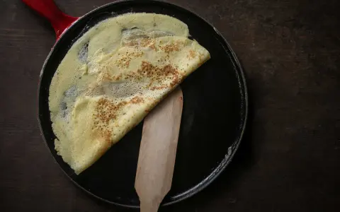 Ein goldbrauner Crêpe wird in einer Pfanne mit rotem Griff gefaltet. Das **Crêpes Teig Rezept Original** gelingt so perfekt.