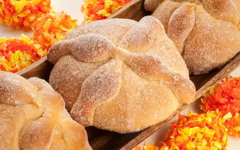 Pan de muerto, süßes Brot mit Zucker bestreut, liegt in einer Holzschale, umgeben von bunten Papierblumen.