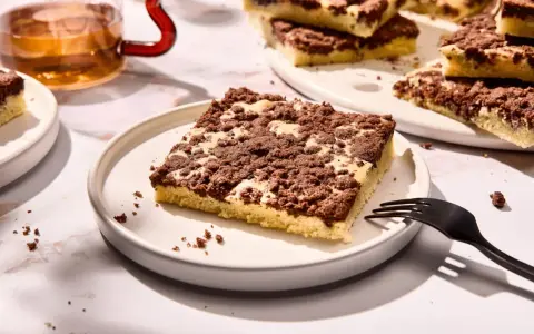 Ein Stück versunkener Streuselkuchen mit Schokostreuseln auf einem Teller, daneben eine Gabel und ein Glas Tee.