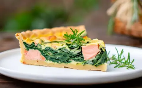 Ein Stück Lachs Spinat Quiche mit Rosmarin garniert auf einem weißen Teller.