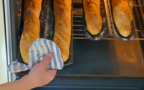 Hand mit Topflappen holt goldbraune Baguettes aus dem Ofen. Der Duft von frisch gebackenen Baguettes liegt in der Luft.