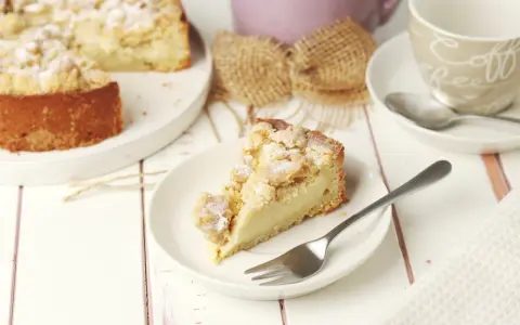 Ein Stück Streuselkuchen mit Pudding auf einem Teller, daneben eine Kaffeetasse und ein ganzer Kuchen.