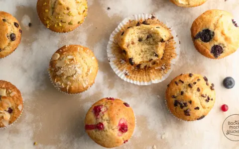 Verschiedene Muffins, darunter Schoko- und Blaubeermuffins. Ein Muffin ist angeknabbert. Die Frage "wie lange muffins backen" wird hier visuell beantwortet: Sie sind perfekt goldbraun.