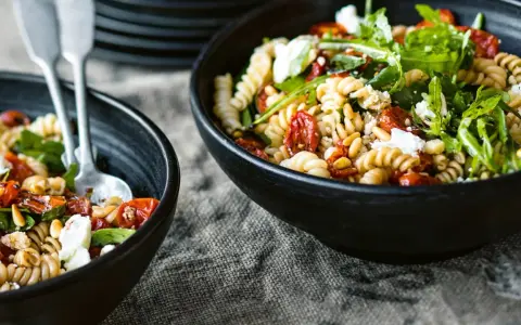 Zwei Schalen mit einem köstlichen nudelsalat mit getrockneten tomaten, garniert mit Rucola und Pinienkernen.