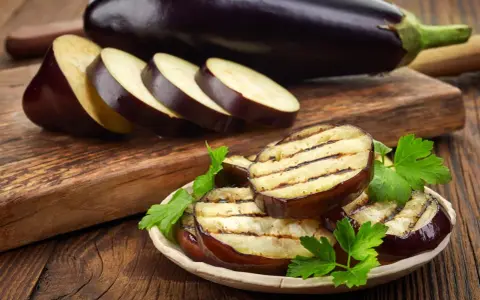 Gegrillte Auberginenscheiben mit Schale, garniert mit Petersilie, liegen auf einem Teller. Daneben liegen weitere Scheiben und eine ganze Aubergine.