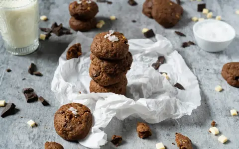 Gestapelte triple chocolate cookies mit Meersalzflocken, umgeben von Schokoladenstückchen und einem Glas Milch.