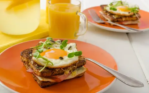 Ein köstlicher Croque Madame mit Spiegelei, Schinken und Käse auf einem orangefarbenen Teller, daneben Saft und Wasser mit Zitrone.