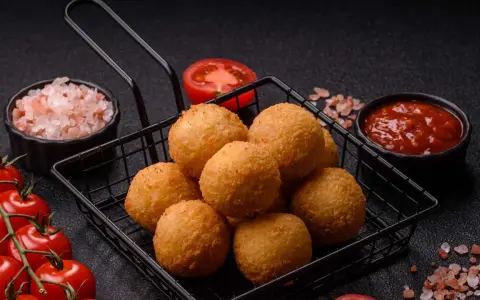 Goldbraune Arancini im Korb, bereit für ein köstliches arancini rezept. Dazu frische Tomaten, Salz und Dip.