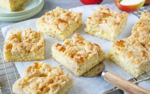 Saftiger Apfelkuchen vom Blech mit knuspriger Streuselhaube. Ein Stück wird gerade mit einem Kuchenheber herausgenommen.