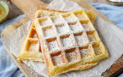 Zwei goldbraune belgische Waffeln, bestäubt mit Puderzucker, liegen auf Backpapier auf einem Holzbrett.