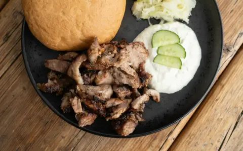 Ein Teller mit gegrilltem Fleisch, Tzatziki, Gurkenscheiben, Krautsalat und einem Brötchen – perfekt für ein leckeres Gyros Rezept.