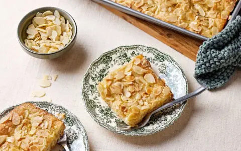Ein Stück Butterkuchen mit Mandelblättchen auf einem Teller, daneben eine Schüssel mit Mandeln. Perfekt für ein Butterkuchen Rezept.