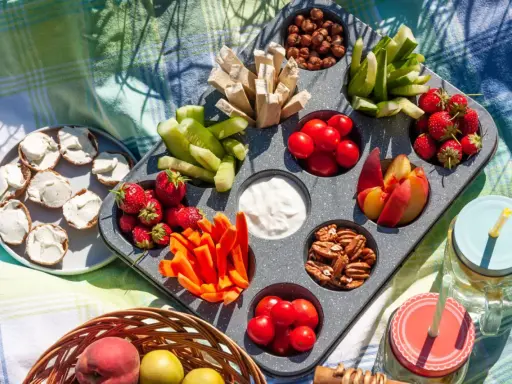 Leckere Picknick-Snacks: Erdbeeren, Gurken, Karotten, Nüsse, Tomaten, Pfirsiche und Brotscheiben mit Frischkäse auf einer Decke.