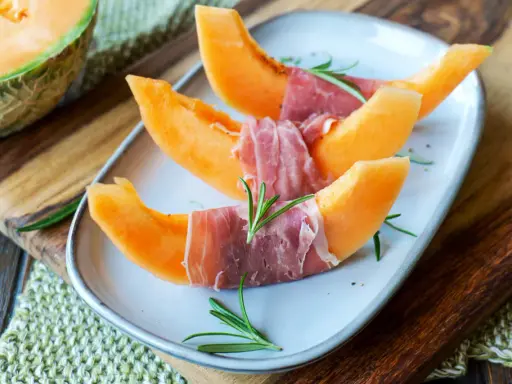 Melone mit Schinken, garniert mit Rosmarin, auf einem Teller angerichtet. Ein halber Melone liegt daneben auf einem Holzbrett.