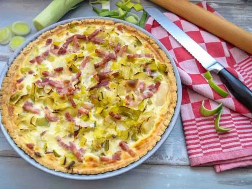 Herzhafte Quiche mit Lauch und Speck, perfekt für herzhafte Quiche Rezepte. Daneben liegen Lauch, ein Messer und ein Nudelholz.