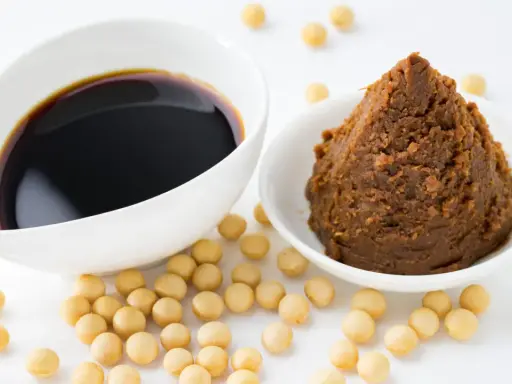 Miso-Paste und Sojasauce in weißen Schalen, umgeben von Sojabohnen. Eine köstliche Alternative zu Hefeflocken für herzhafte Gerichte.