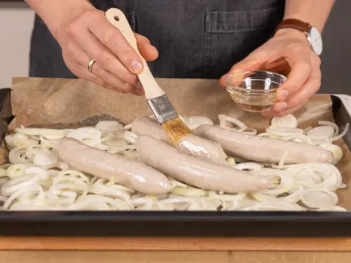 Bratwurst im Backofen: Zwiebelringe auf Blech, darauf vier Würste. Mit Pinsel wird eine Wurst mit Flüssigkeit bestrichen.