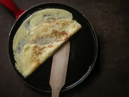 Ein goldbrauner Crêpe wird in einer Pfanne mit rotem Griff gefaltet. Das **Crêpes Teig Rezept Original** gelingt so perfekt.