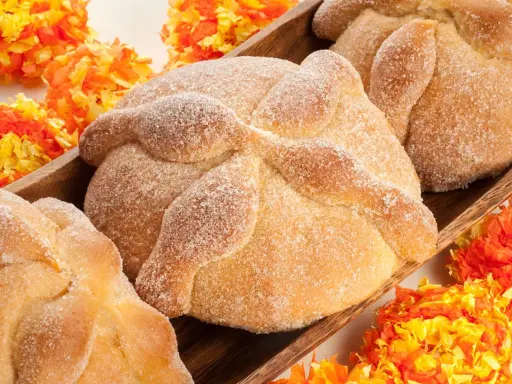 Pan de muerto, süßes Brot mit Zucker bestreut, liegt in einer Holzschale, umgeben von bunten Papierblumen.