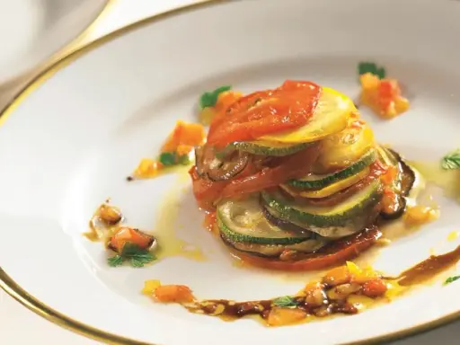 Ein kunstvoll geschichtetes Ratatouille-Rezept, inspiriert vom Film, mit Tomaten, Zucchini und Aubergine auf einem Teller mit goldenem Rand.