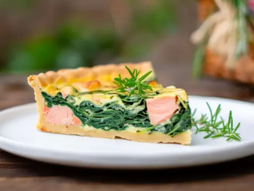 Ein Stück Lachs Spinat Quiche mit Rosmarin garniert auf einem weißen Teller.
