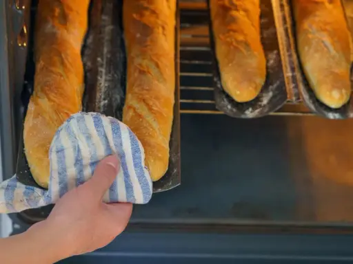 Hand mit Topflappen holt goldbraune Baguettes aus dem Ofen. Der Duft von frisch gebackenen Baguettes liegt in der Luft.