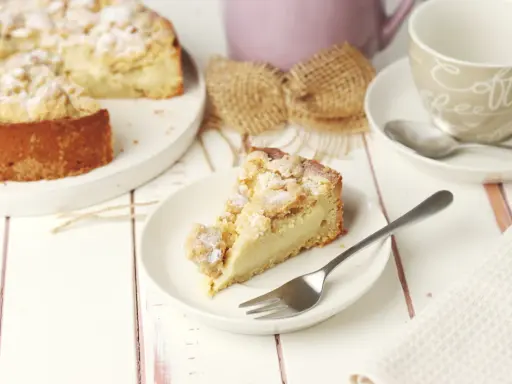Ein Stück Streuselkuchen mit Pudding auf einem Teller, daneben eine Kaffeetasse und ein ganzer Kuchen.