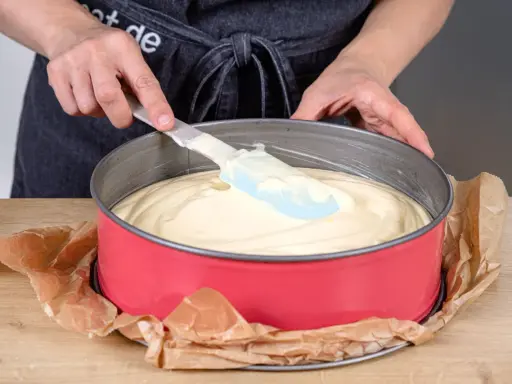 Hände verteilen den Teig für einen Joghurtkuchen mit 500 g Joghurt in einer roten Springform.