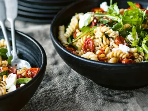 Zwei Schalen mit einem köstlichen nudelsalat mit getrockneten tomaten, garniert mit Rucola und Pinienkernen.