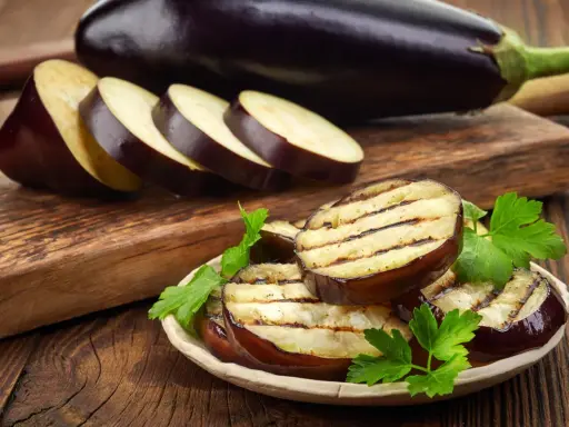 Gegrillte Auberginenscheiben mit Schale, garniert mit Petersilie, liegen auf einem Teller. Daneben liegen weitere Scheiben und eine ganze Aubergine.