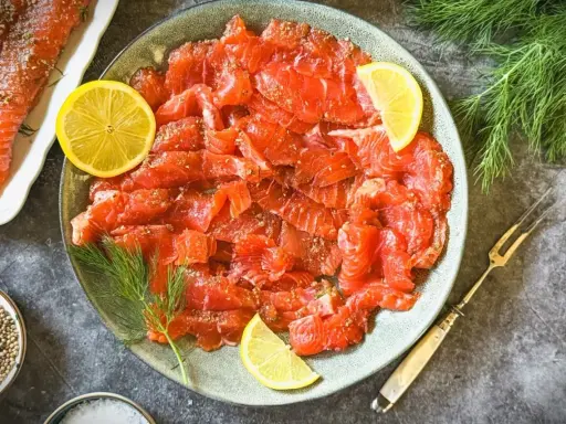Frisch gebeizter Lachs auf einem Teller, garniert mit Zitronenscheiben und Dill. Daneben eine Schale mit Koriandersamen.