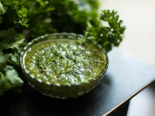 Frische Kräuter umgeben eine Schale mit leuchtend grünem Chimichurri. Was ist Chimichurri? Eine würzige argentinische Sauce aus Petersilie, Knoblauch und Essig.