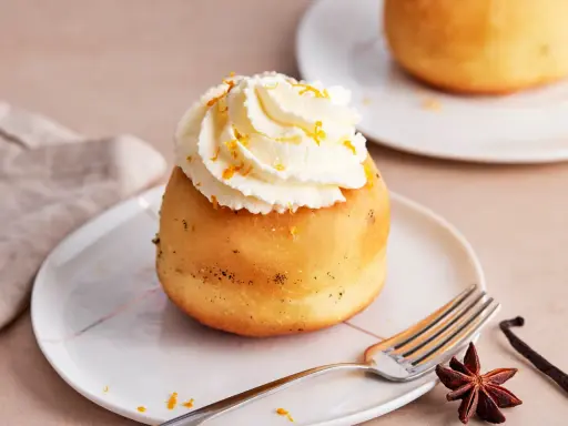 Ein köstlicher Baba au Rhum, gekrönt mit Schlagsahne und Orangenzesten, wartet auf einem Teller. Daneben liegen Sternanis und Vanilleschote.