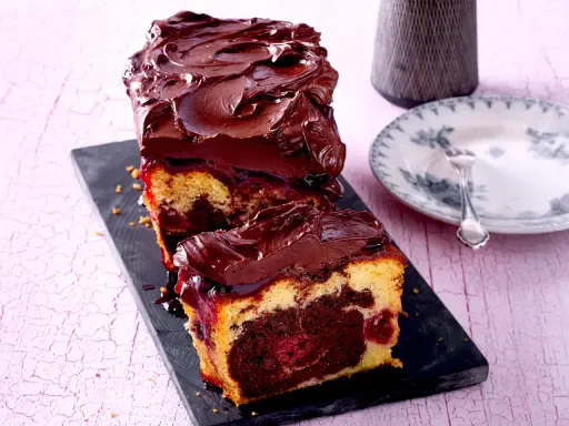 Saftiger Marmorkuchen mit Kirschen, überzogen mit dunkler Schokoladenglasur. Ein Stück ist bereits angeschnitten und zeigt die köstliche Marmorierung.