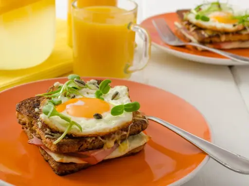 Ein köstlicher Croque Madame mit Spiegelei, Schinken und Käse auf einem orangefarbenen Teller, daneben Saft und Wasser mit Zitrone.