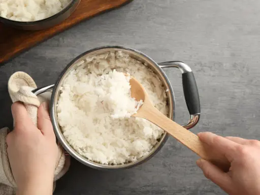 Hände halten einen Topf mit frisch gekochtem Reis. Eine Holzschaufel hebt eine Portion Reis heraus. Die Frage "wie lange kocht man reis" wird hier visuell beantwortet.