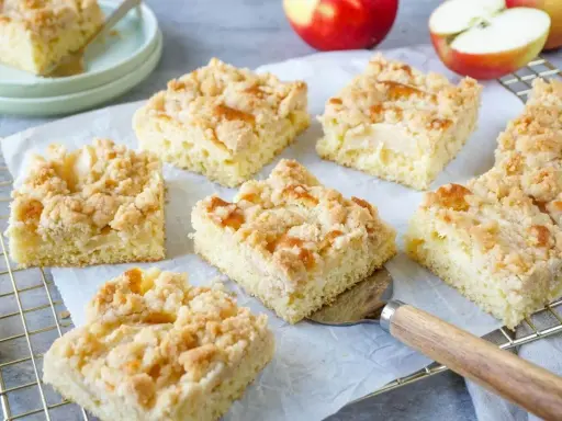 Saftiger Apfelkuchen vom Blech mit knuspriger Streuselhaube. Ein Stück wird gerade mit einem Kuchenheber herausgenommen.