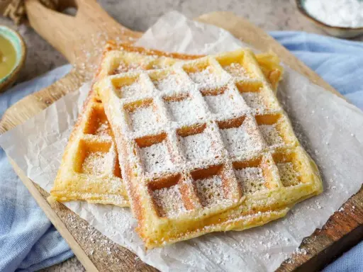 Zwei goldbraune belgische Waffeln, bestäubt mit Puderzucker, liegen auf Backpapier auf einem Holzbrett.