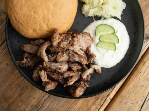 Ein Teller mit gegrilltem Fleisch, Tzatziki, Gurkenscheiben, Krautsalat und einem Brötchen – perfekt für ein leckeres Gyros Rezept.