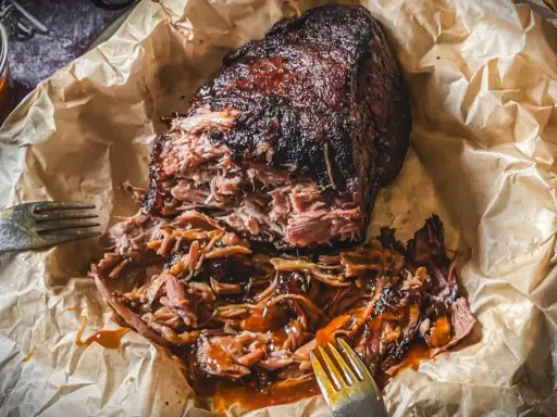 Saftiges pulled pork, zart zerfallen, mit BBQ-Sauce übergossen, auf Backpapier mit Gabeln.