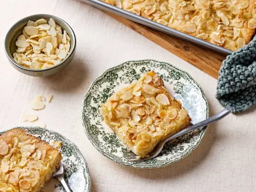 Ein Stück Butterkuchen mit Mandelblättchen auf einem Teller, daneben eine Schüssel mit Mandeln. Perfekt für ein Butterkuchen Rezept.