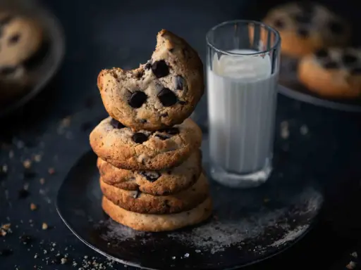 Ein Stapel amerikanischer Cookies mit Schokoladenstückchen, daneben ein Glas Milch. Ein Cookie ist angeknabbert.