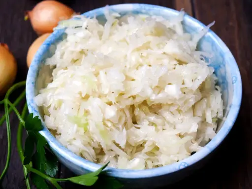 Schüssel mit frisch zubereitetem Krautsalat, daneben Zwiebeln und Petersilie. Perfekt, um Krautsalat selbst zu machen!